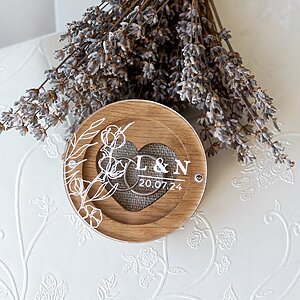 Round wooden ring box with a clear acrylic lid, decorated with floral engraving, initials, and a date. Inside is a heart-shaped slot for wedding rings