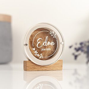 Round white plaster pet fur keepsake box with a floral wreath design on the lid named Edna, standing on an oak block with a reflection on a white table.