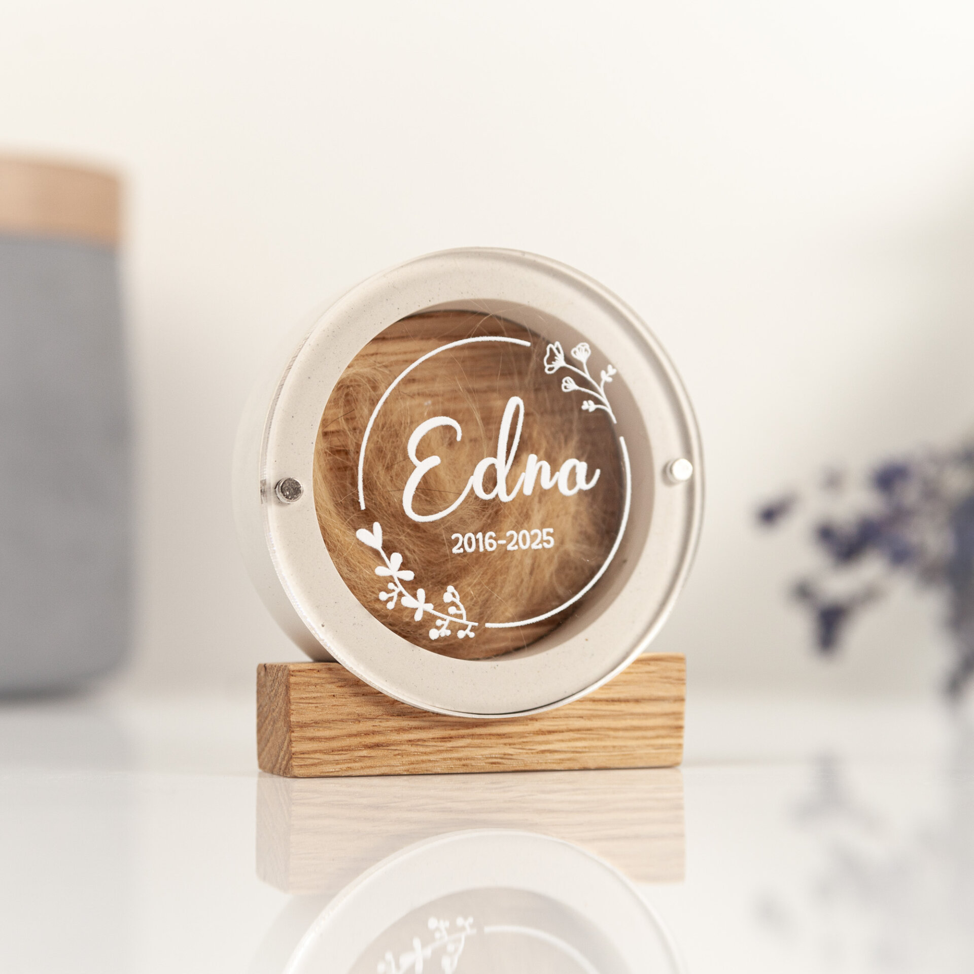 Round white plaster pet fur keepsake box with a floral wreath design on the lid named Edna, standing on an oak block with a reflection on a white table.